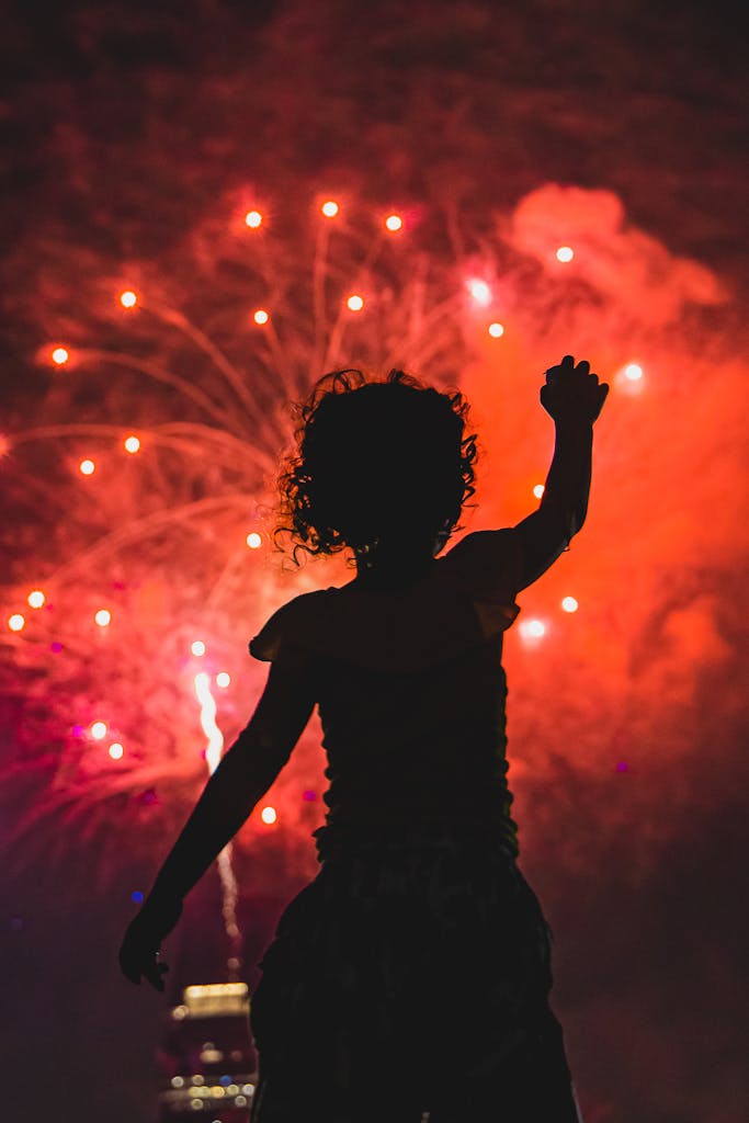 Silhouette of a child raising a hand under a vivid fireworks show lights up the night sky.